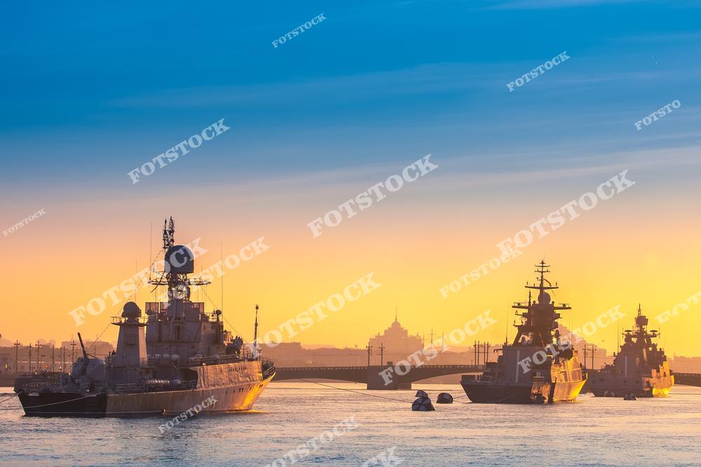 На фотографии изображены военные корабли, стоящие на реке Неве в Санкт-Петербурге. На заднем плане виден силуэт зданий и мостов города. Рассветные лучи создают атмосферу торжественности и гармонии. Гладкая поверхность воды подчёркивает спокойствие утреннего Петербурга.
